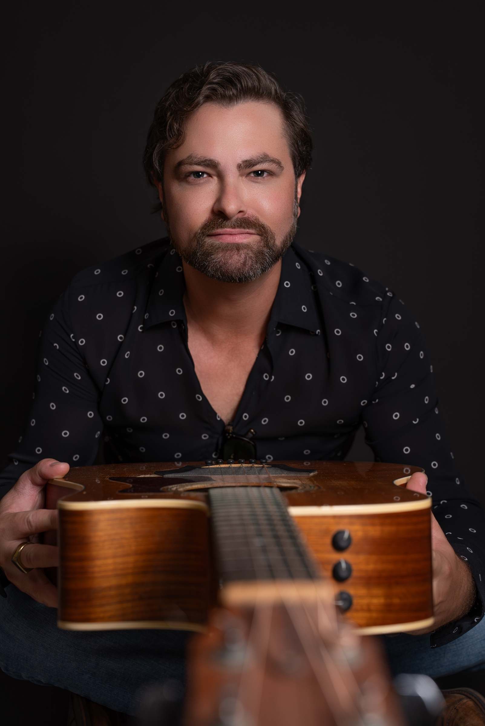 Portrait of Willy B looking into the camera, holding his acoustic guitar toward the lens